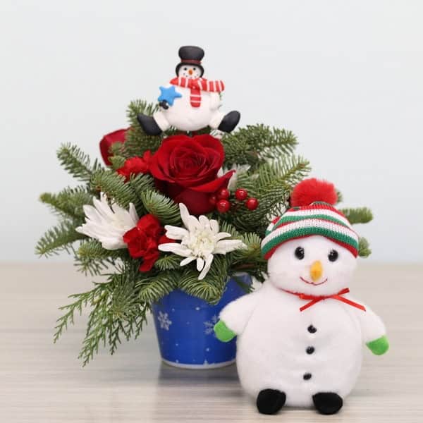 Arrangement floral de Noël Mini Sweets dans un pot bleu avec décoration bonhomme de neige, accompagné d’une peluche bonhomme de neige assortie.