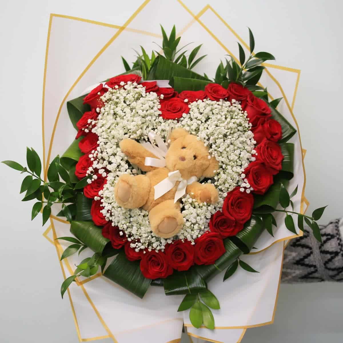 Heart-shaped bouquet of 24 red roses with baby’s breath and a teddy bear in the center.