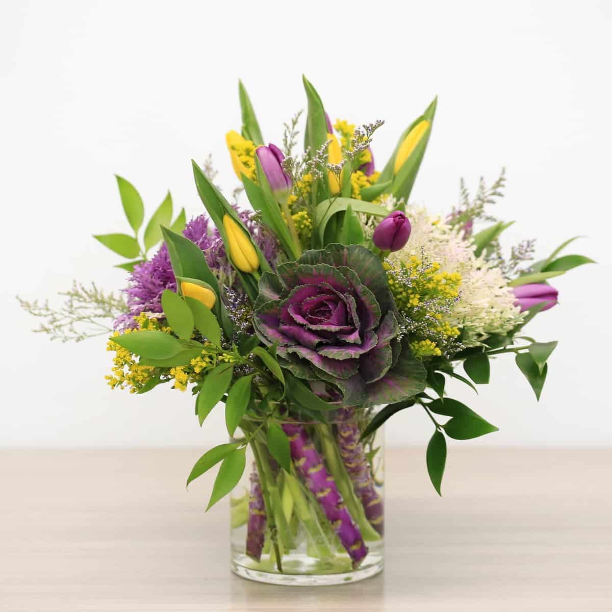 Violet Embrace arrangement with decorative kale, purple and yellow tulips, solidago and caspia in etched Le Bouquet vase.